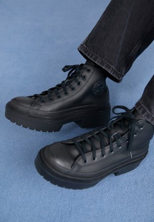 Black leather high-top boots with chunky soles worn with dark pants on a light blue textured surface.