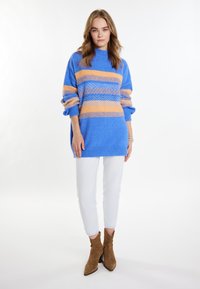 Maglione blu lavorato a maglia con righe arancioni, dotato di un collo alto, vestibilità rilassata e maniche a palloncino. Abbinato a pantaloni bianchi e stivaletti marroni.