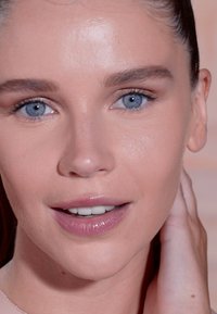 Close-up van het gezicht van een persoon met steil bruin haar. Kenmerken zijn een heldere huid, blauwe ogen, natuurlijke make-up en een subtiele lipglans.