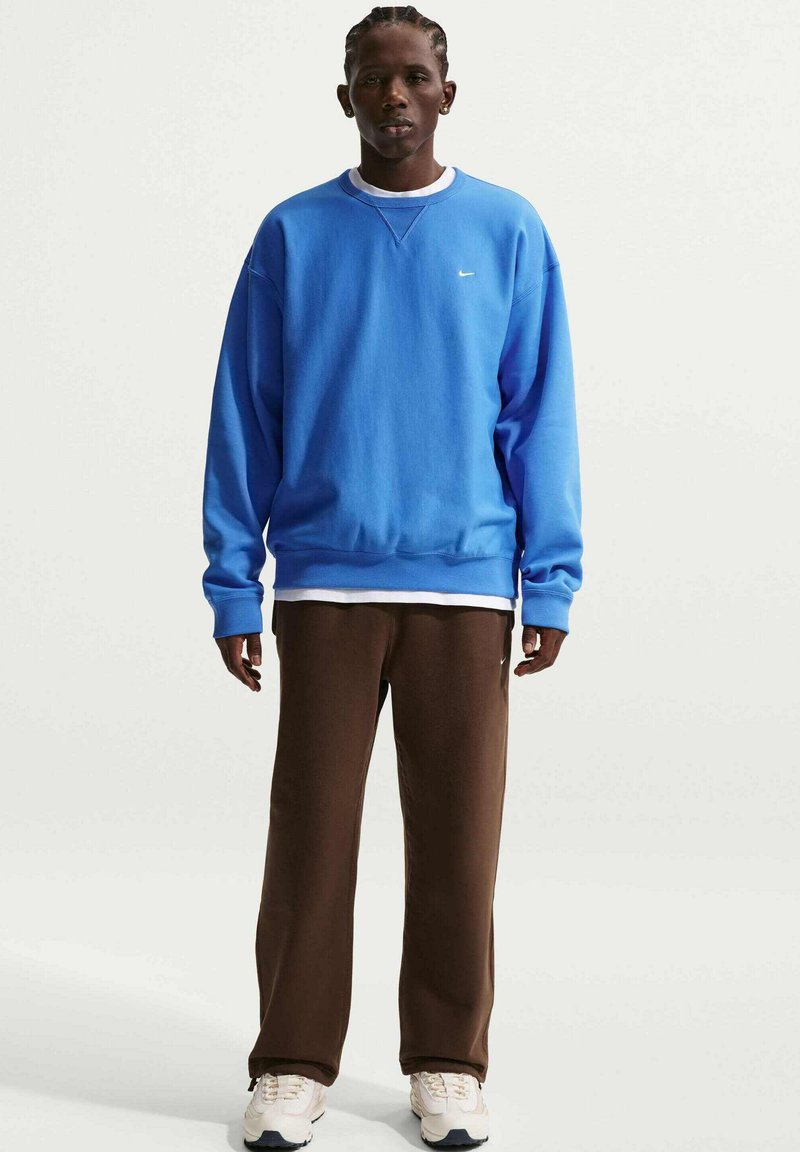 Mann steht nach vorne blickend, trägt einen blauen Pullover, weißes Unterhemd, braune Hose und weiße Turnschuhe vor einem schlichten weißen Hintergrund.