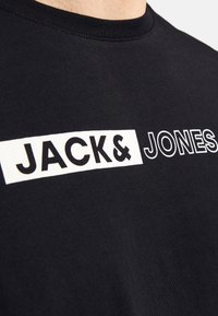 Schwarzes T-Shirt mit kurzen Ärmeln und Rundhalsausschnitt. Verfügt über weiße Blockbuchstaben mit der Aufschrift "JACK & JONES" über der Brust. Weicher Stoff.