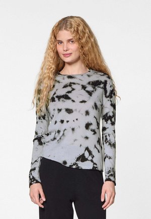 Blonde Frau mit langen, lockigen Haaren, die ein graues und schwarzes Batik-Langarmshirt und eine schwarze Hose trägt, vor einem schlichten Hintergrund stehend.