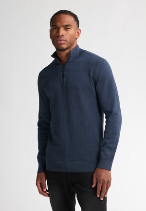 Marineblauw zip-up sweater met textuurpatroon, hoge kraag en geribbelde boorden. Gemaakt met zwarte jeans, tegen een effen witte achtergrond.