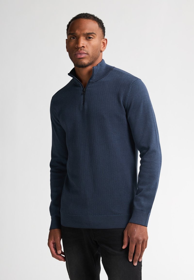 Felpa zip-up blu navy con motivo testurizzato, colletto alto e polsini a costine. Indossata con jeans neri, su uno sfondo bianco semplice.