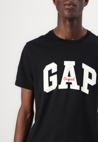 Fekete pamut póló, amelyen vastag fehér betűkkel szerepel a "GAP" felirat, alatta pedig kisebb piros betűkkel az "Original" szó. Rövid ujjak, kerek nyakkivágás.