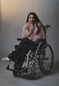 Jeune femme en fauteuil roulant portant un manteau en fausse fourrure rose, un pantalon noir pailleté et des chaussures argentées scintillantes, posant devant un fond neutre.