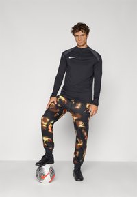 Black athletic long-sleeve top with a zipper, gray accents, and black patterned joggers in orange and yellow tones, paired with a soccer ball.