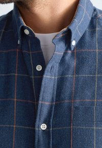 Camicia di flanella blu con un motivo a quadri giallo e arancione, caratterizzata da un colletto button-down e bottoni bianchi. Include una tasca sul petto.