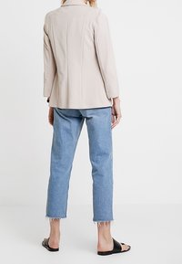 Blazer beige sur mesure avec épaules structurées, associé à un jean bleu clair court aux ourlets effilochés et des sandales plates noires.