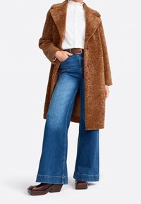 Cappotto marrone con texture, camicia bianca con colletto, jeans a zampa d'elefante blu e décolleté marroni con zeppa. Il cappotto ha grandi bottoni e un ampio rever.