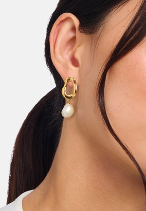 Orecchino d'oro con una forma aperta irregolare e una perlina bianca pendente, indossato all'orecchio di una donna con capelli scuri raccolti.