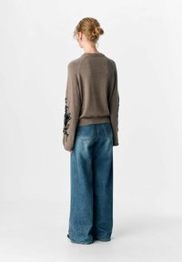 Frau mit einem Dutt trägt einen braunen Pullover mit schwarzer Blumenstickerei an den Ärmeln und weit geschnittenen blauen Jeans, steht vor einem weißen Hintergrund.