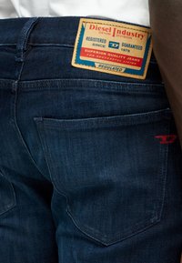 Σκούρα μπλε τζιν παντελόνι με τσέπη, στην οποία είναι ραμμένο ένα μπεζ και κόκκινο λογότυπο της Diesel Industry, πάνω από αυτήν, στη μέση ενός ατόμου.