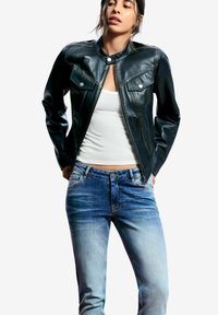 Chaqueta de cuero negra con cuello alto, cierre de cremallera y dos bolsillos en el pecho. Llevada sobre una camiseta sin mangas blanca y jeans azul claro desgastados.