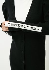 Schwarze Strickjacke mit tiefem V-Ausschnitt, gerippten Bündchen und weißen Ärmelakzenten mit schwarzen geometrischen Mustern. Fünf Knöpfe vorne.