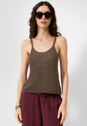 Vrouw die zwarte cat-eye zonnebril, een bruin gehaakt tanktopje en losse bordeauxrode broek met een trekkoord in de taille draagt.