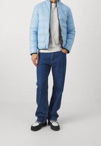 Ljusblå puffjacka med ståkrage, svart dragkedja, grå t-shirt under, kombinerad med mörkblå jeans och svart-vita sneakers.