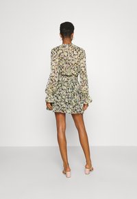 Donna in piedi, di spalle, vestita con un mini abito a strati a maniche lunghe con fantasie floreali e sandali con tacco a blocco rosa, su uno sfondo neutro.