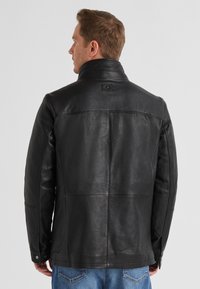 Schwarze Lederjacke mit hohem Kragen, sichtbaren Nähten und glatter Textur. Verfügt über knöpfbare Manschetten und ein Logo-Detail auf der Rückseite.