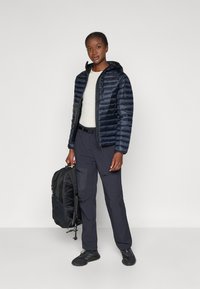 Veste matelassée bleu marine avec capuche, en matériau léger, associée à un pantalon cargo foncé et un sac à dos, avec des poches zippées et des baskets noires.