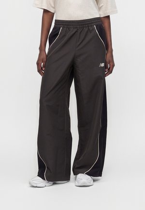 Person iført sorte New Balance trackpants med vide ben og hvide pibelinjer samt hvide sneakers på en ensfarvet baggrund.