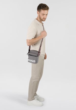 Borsa a tracolla grigia con cerniera nera e logo "HUGO" bianco, dotata di una tracolla regolabile decorata con un motivo rosso e nero. Design rettangolare compatto.