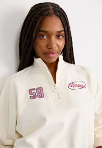 Felpa a collo alto di colore crema con zip parziale, caratterizzata da un ricamo del numero "59" in rosso e rosa, con il logo "ellesse" in evidenza sul petto. Tessuto morbido.