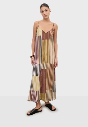 Kobieta stojąca w bezrękawnej, wielokolorowej sukience maxi w paski i czarnych sandałach z odkrytymi palcami na jasnym, jednolitym tle.