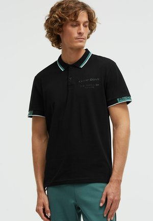Mann mit lockigem Haar, der ein schwarzes Poloshirt mit grün-weißem Besatz und türkisfarbene Hose trägt, schaut nach unten und zur Seite vor einem einfarbigen Hintergrund.