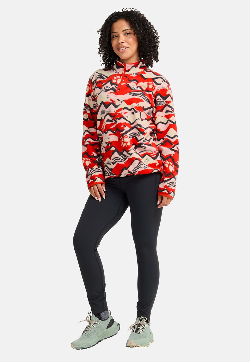 Femme debout, mains détendues, portant une veste polaire rouge à motifs, un legging noir, des chaussettes grises et des baskets vertes sur fond blanc.