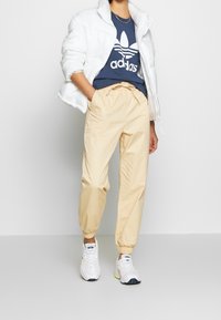 Bílá puffová bunda s vysokým límcem, námořnické tričko Adidas s bílým logem a béžové tepláky s elastickým pasem, doplněné bílými teniskami.