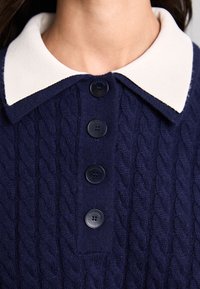 Marinblå kabelstickad tröja med en vit kontrasterande krage, fyra mörka knappar och texturerad stickad mönster.