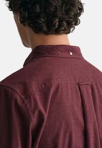 Camisa burdeos con cuello, de tejido texturizado, que presenta un pliegue en la parte posterior y un detalle decorativo de botón a presión en el cuello.