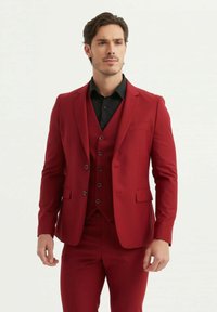 Costume rouge ajusté avec une veste à revers cranté, un gilet assorti et un pantalon slim. Le tissu semble lisse, avec une fermeture à deux boutons à l'avant.