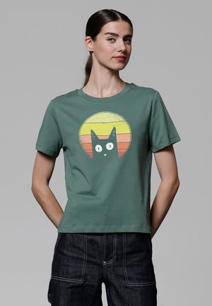 T-shirt en coton vert avec un col rond, présentant un graphique d'un chat noir sur fond de coucher de soleil dans des teintes jaunes et oranges.
