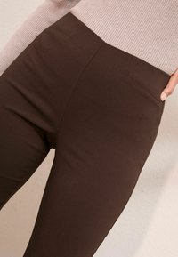 Közelkép egy személyről, aki magas derekú, testhezálló, sötétbarna nadrágot visel, oldalsó és hátsó zsebekkel, amit egy világos rózsaszín, bordázott felsővel párosított.