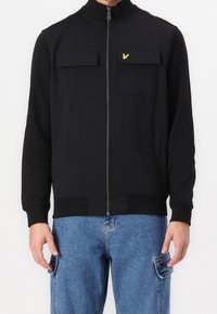 Chaqueta negra con cremallera y cuello alto, dos bolsillos frontales y una cinturilla ajustada; hecha de un tejido suave con un pequeño logo amarillo.