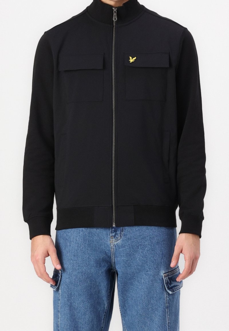 Chaqueta negra con cremallera y cuello alto, dos bolsillos frontales y una cinturilla ajustada; hecha de un tejido suave con un pequeño logo amarillo.