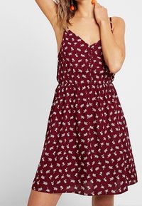 Burgundy sommarklänning med vitt blommönster, V-ringning, justerbara spaghettiband och elastisk midja för en avslappnad passform.