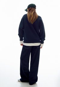 Námořnická oversized mikina vrstvená přes bílé tričko s dlouhým rukávem, doplněná širokými tmavými džíny. Zelená čepice podtrhuje celý vzhled.