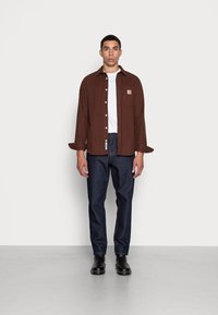 Bruine lange-mouwen overhemd met knopen en borstzak, gedragen over een wit T-shirt, gecombineerd met donkerblauwe jeans en zwarte laarzen. Effen achtergrond.