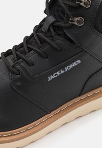 Zapatilla alta sintética negra con un acabado texturizado, detalles de malla, ojales de metal y una suela de color beige en contraste. Logo "JACK&JONES" exhibido.