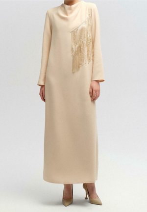 Robe beige à manches longues avec un col montant et un détail de franges en cascade sur la poitrine, associée à des talons beiges à bouts pointus.