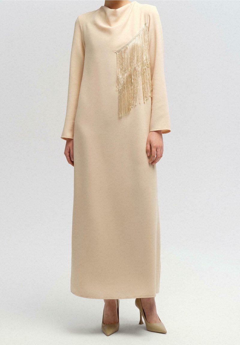Robe beige à manches longues avec un col montant et un détail de franges en cascade sur la poitrine, associée à des talons beiges à bouts pointus.