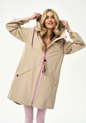 Kobieta ubrana w beżową pelerynę przeciwdeszczową z kapturem, różowym zamkiem błyskawicznym i kolorowymi frędzlami, trzymająca kaptur obiema rękami, uśmiechająca się, na białym tle.