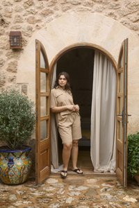 Femme en short beige et chemise, debout dans une ouverture de porte voûtée en bois avec un mur en pierre et de grandes jardinières de chaque côté.