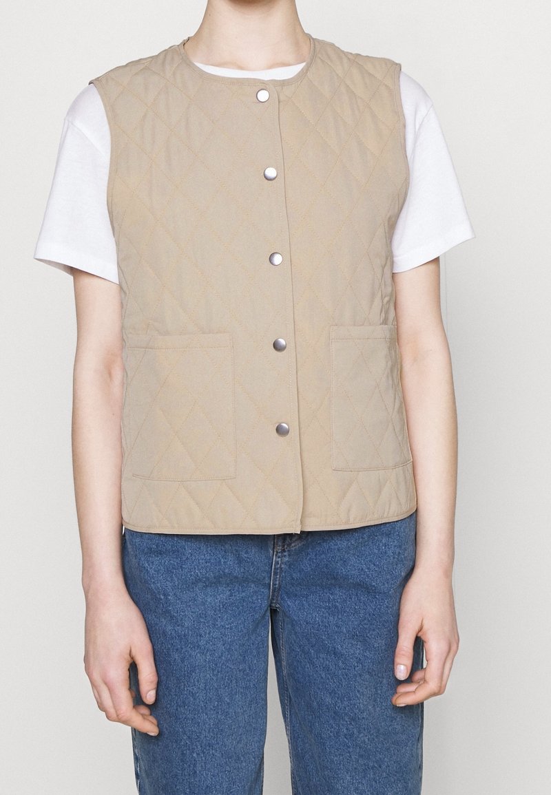 Veste matelassée beige avec boutons-pression et poches avant portée sur un t-shirt blanc à manches courtes, associée à un jean bleu.