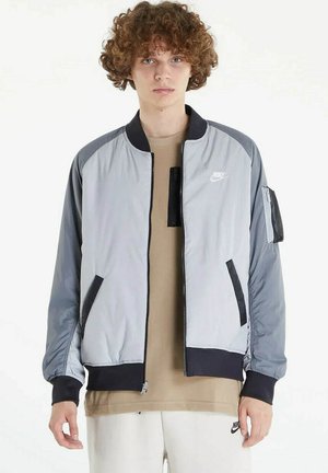 Giovane con capelli ricci che indossa una giacca Nike con cerniera grigio chiaro e nero sopra una maglietta beige e pantaloni bianchi Nike.