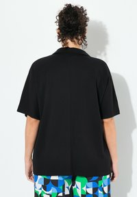 Černá oversized blůza s límečkem a krátkými rukávy, s rovnou zadní švem. Tkanina vypadá hladce a lehce.