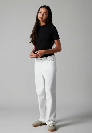 Młoda kobieta o długich ciemnych włosach, ubrana w czarną, krótko rękawową bluzkę, białe dżinsy i beżowe trampki, stojąca na tle jednolitego szarego tła.